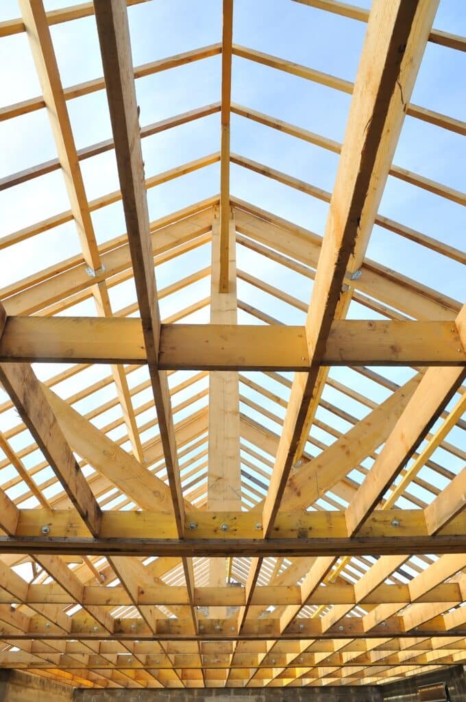 Charpente bois neuve : Ossature de toit en structure porteuse Structure de charpente en bois neuve sous un ciel bleu clair, vue en contre-plongée montrant les fermes de toit et les chevrons assemblés.