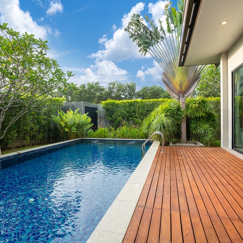Piscine rectangulaire privée avec mosaïque bleue et terrasse en bois, entourée de végétation luxuriante sous un ciel bleu nuageux.
