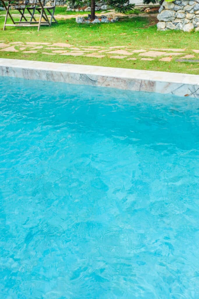 Piscine Turquoise avec Bordure en Marbre et Jardin Eau turquoise agitée d'une piscine bordée de marbre, avec une pelouse et des meubles de jardin en arrière-plan.